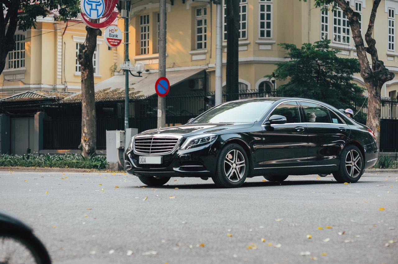 A sleek black sedan navigates an urban street lined with historic buildings and lush trees.